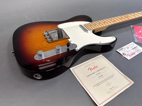 Fender electric guitar in sunburst finish with maple neck and chrome hardware alongside Fender certificate of authenticity and tags on gray background