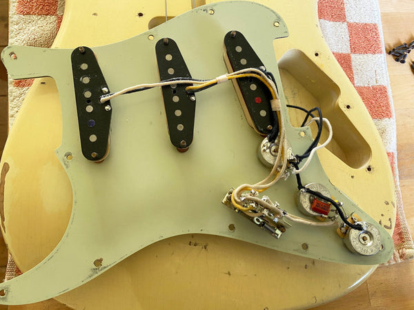 Vintage guitar pickguard with three single-coil pickups and wiring, set on a worn cream-colored guitar body, showing aged components and potentiometers.