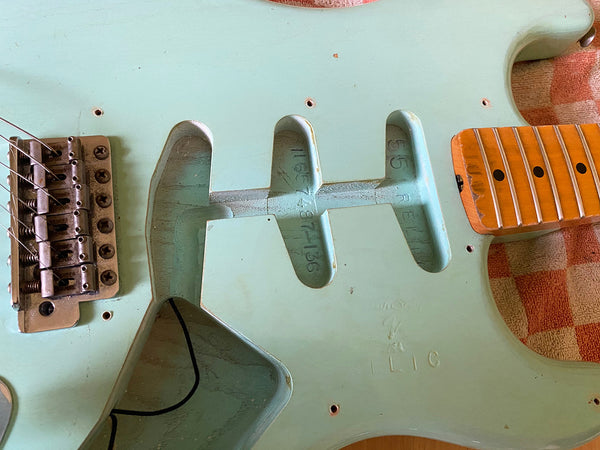 Close-up of vintage seafoam green electric guitar body with pickup cavities, tremolo bridge, and maple neck with fretboard dots visible on a checkered cloth surface.