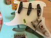 Close-up of light blue electric guitar body with white pickguard removed, showing three single-coil pickups, wiring, potentiometers, and vintage-style bridge.
