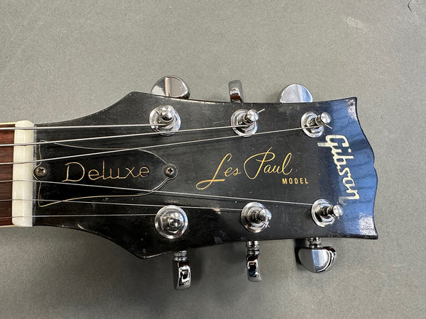 Close-up of Gibson Les Paul Deluxe electric guitar headstock showing tuning pegs, logo, and strings on gray background