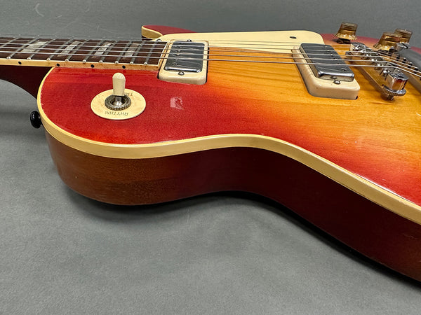 Close-up of cherry sunburst electric guitar body with cream binding, chrome pickups, and rhythm/treble toggle switch on gray surface