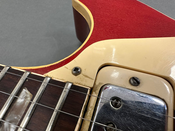 Close-up of vintage red electric guitar body showing neck joint, cream pickguard, metal pickup cover, and fretboard with strings and inlays