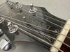 Close-up of Deluxe model guitar headstock showing tuning pegs, strings, and logo plate on black finish wood.