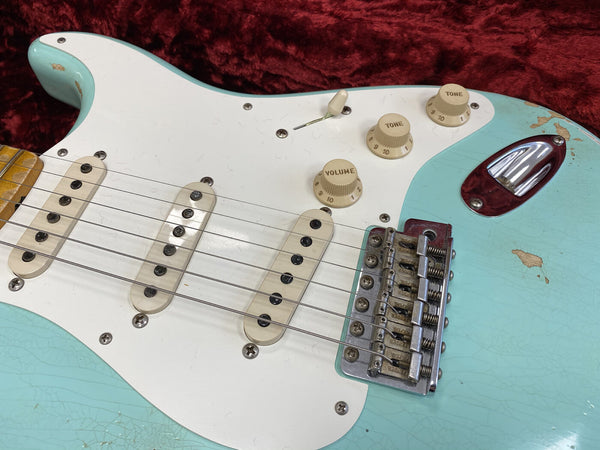 Close-up of vintage blue electric guitar body with white pickguard, three single-coil pickups, tone and volume knobs, and chrome bridge on red plush background