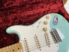 Close-up of vintage light blue electric guitar body with white pickguard, three single-coil pickups, tone and volume knobs, and maple neck on red plush case lining