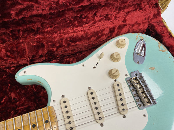 Close-up of vintage light blue electric guitar body with white pickguard, three single-coil pickups, tone and volume knobs, and maple neck on red plush case lining