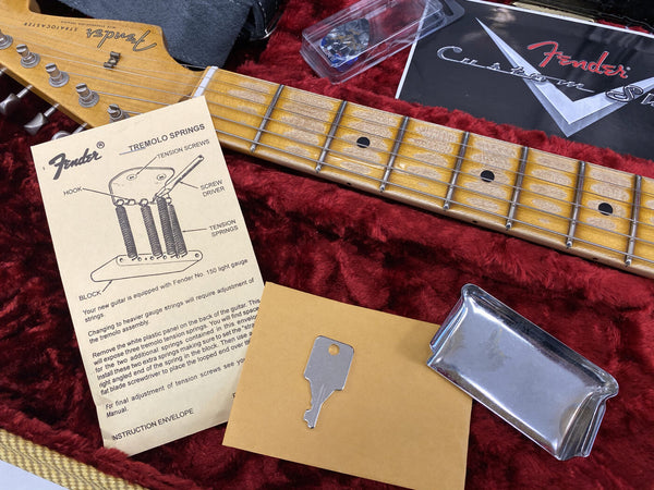 Close-up of Fender Stratocaster guitar neck with vintage maple fretboard, accompanying Fender tremolo springs instruction sheet, metal key on envelope, chrome pickup cover, and Fender Custom Shop case lining in red plush material.