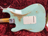 Close-up of the back of a worn light blue Fender Stratocaster electric guitar with a maple neck, showing paint chips and a white backplate, resting on a red velvet guitar case interior.