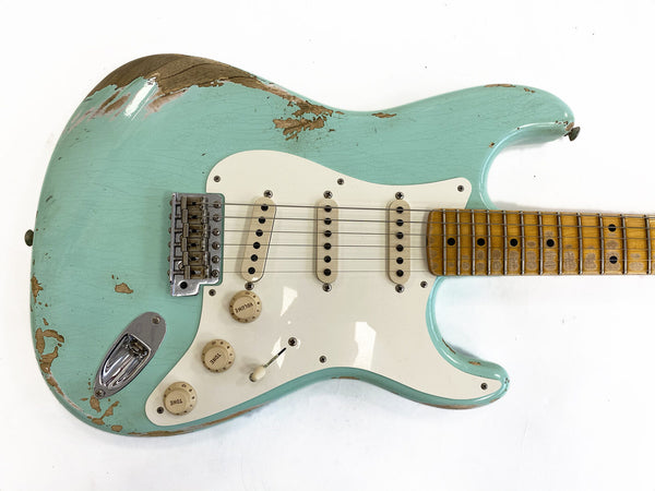 Close-up of a vintage light blue relic electric guitar with three single-coil pickups, worn wood finish, white pickguard, and maple neck with dot inlays against a white background.