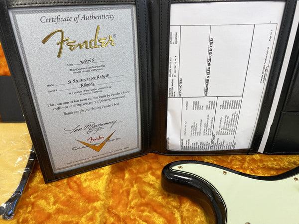 Certificate of Authenticity for Fender 61 Stratocaster Relic with Fender Custom Shop specifications and electronic hardware notes in black binder on yellow plush surface next to black and white guitar body.