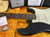 Close-up of black Fender Stratocaster electric guitar with rosewood fretboard, white pickguard, and three single-coil pickups on a yellow plush guitar case interior, showing Fender Custom Shop certificate and accessories including guitar picks and inspection card.