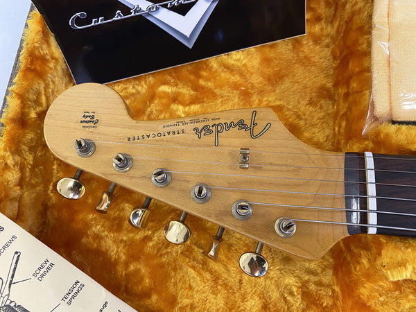 Close-up of Fender Stratocaster guitar headstock with vintage tuners in orange plush-lined case, showing logo and string details for classic electric guitar maintenance and repair.