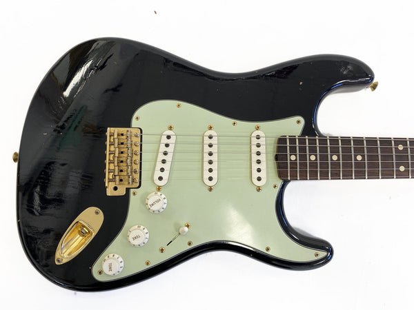 Black electric guitar with mint green pickguard, three single-coil pickups, volume and tone control knobs, and gold hardware on white background