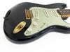 Close-up of black electric guitar body with white pickguard, three single-coil pickups, gold hardware, and tone and volume control knobs on white background.