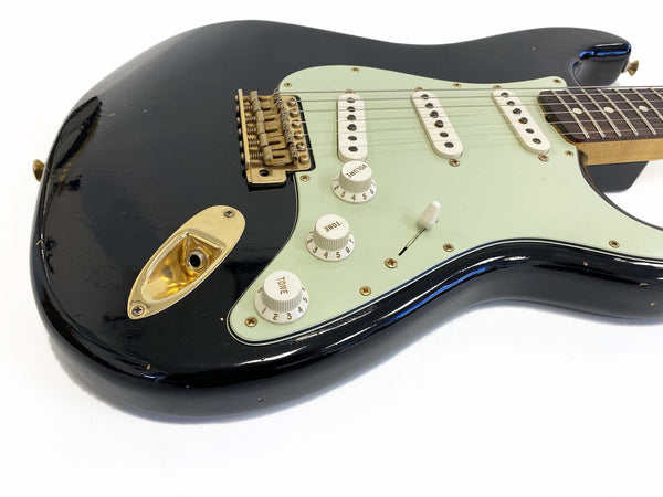 Close-up of black electric guitar body with white pickguard, three single-coil pickups, gold hardware, and tone and volume control knobs on white background.