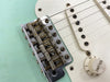 Close-up of vintage Fender electric guitar bridge, strings, white volume knob, and aged cream pickup cover on seafoam green body