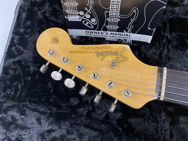 Close-up of Fender Stratocaster guitar headstock with tuning pegs and strings, resting on black plush guitar case lining, owner's manual visible in background.