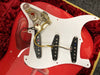 Close-up of disassembled red electric guitar body showing white pickguard with three black single-coil pickups and internal wiring with potentiometers and capacitors exposed, resting on red plush guitar case lining.