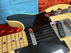 Close-up of vintage-style Telecaster guitar with maple neck, black pickguard, and chrome bridge pickup, resting on a red plush guitar case with blue hardware and electronics notes in the background.