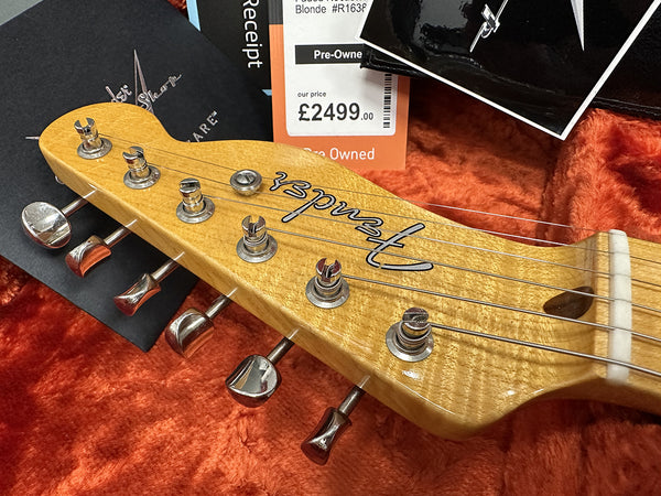 Close-up of Fender guitar headstock with tuning pegs and strings on orange plush background, pre-owned Fender guitar price tag showing £2499, warranty card and receipt in case