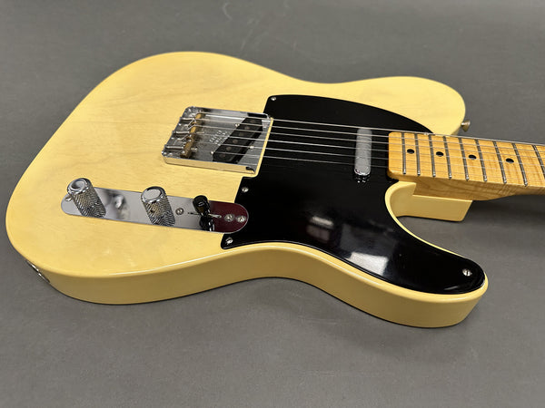 Close-up of vintage-style cream electric guitar body with black pickguard, chrome bridge, and control knobs on gray surface