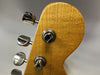 Close-up of natural wood guitar headstock with chrome tuning pegs and strings attached, showing textured finish and tuning mechanisms.