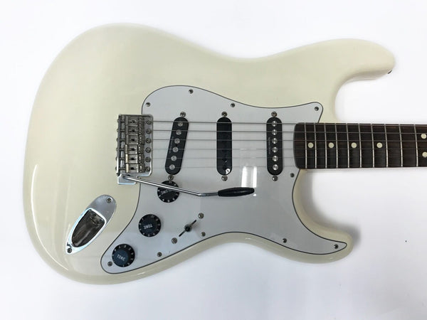 White electric guitar with rosewood fretboard, three single-coil pickups, whammy bar, and black tone and volume knobs on a white background