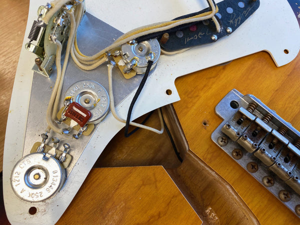 Close-up view of vintage guitar potentiometers, capacitor, wiring and bridge assembly on a natural wood electric guitar body.