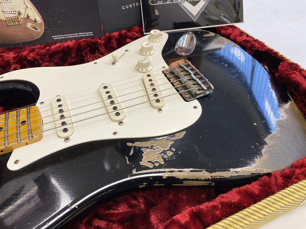 Close-up of a black electric guitar with white pickguard and three single-coil pickups, showing worn and chipped paint on the body, resting in a red velvet-lined guitar case with vintage guitar brochures in the background.