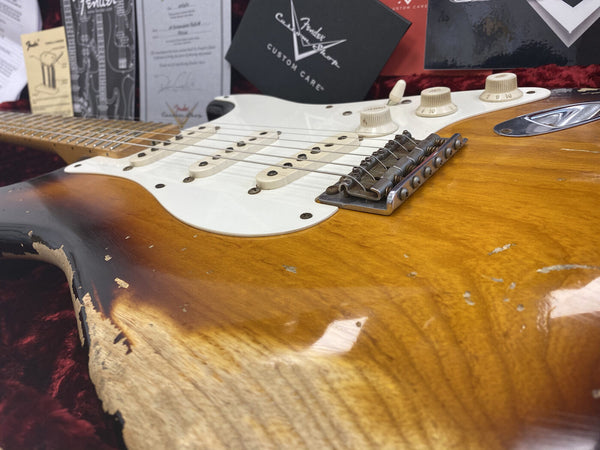 Close-up of a vintage sunburst electric guitar body with worn finish, white pickguard, three single-coil pickups, and white control knobs, displayed on red velvet with Fender Custom Care certificates in the background.