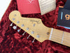 Close-up of Fender Stratocaster guitar headstock with tuning pegs, maple neck, and original custom shop care booklet inside red plush-lined case.