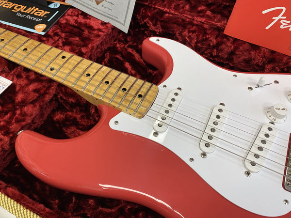 Close-up of red Fender Stratocaster electric guitar with maple neck and white pickguard in plush red guitar case, showing pickups, volume knob, and part of the headstock area with documents and Fender logo visible.