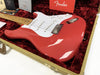 Fender Stratocaster electric guitar in red with white pickguard resting in a plush red-lined tweed hard case, showing maple neck, chrome bridge, and volume and tone knobs, with Fender owner's manual and guitar care documents in the background.