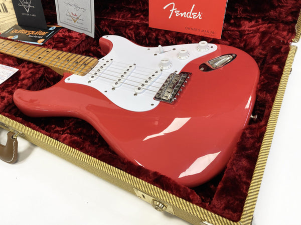 Fender Stratocaster electric guitar in red with white pickguard resting in a plush red-lined tweed hard case, showing maple neck, chrome bridge, and volume and tone knobs, with Fender owner's manual and guitar care documents in the background.