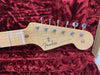 Close-up of Fender Stratocaster guitar headstock with tuning pegs and logo on natural wood finish, placed on red velvet guitar case interior