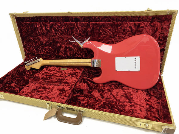 Back of coral red Fender electric guitar with maple neck resting inside an open vintage-style tweed case lined with plush red velvet
