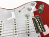 Close-up of red electric guitar body with white pickguard, three single-coil pickups, volume and tone control knobs, and chrome bridge Assembly