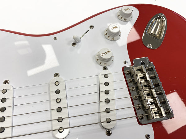 Close-up of red electric guitar body with white pickguard, three single-coil pickups, volume and tone control knobs, and chrome bridge Assembly