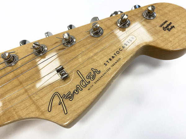 Close-up of natural wood Fender Stratocaster guitar headstock with tuning pegs and strings, featuring original contour body label and synchronized tremolo text
