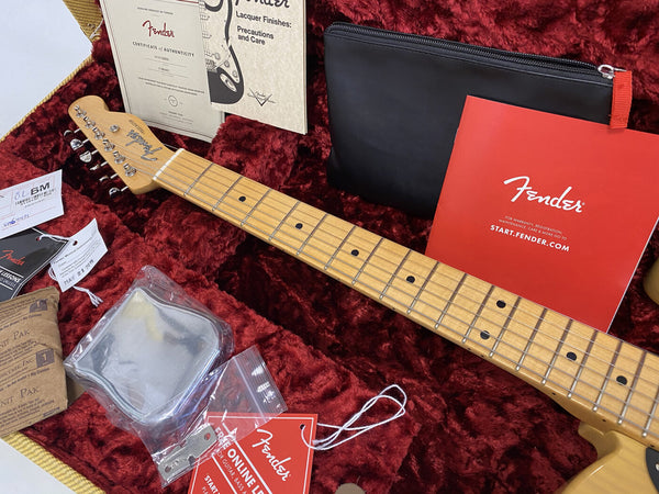 Close-up of Fender guitar neck in plush red case with Fender certificate of authenticity, black zipped pouch, red warranty registration booklet, string pack, tag, and online lessons card.