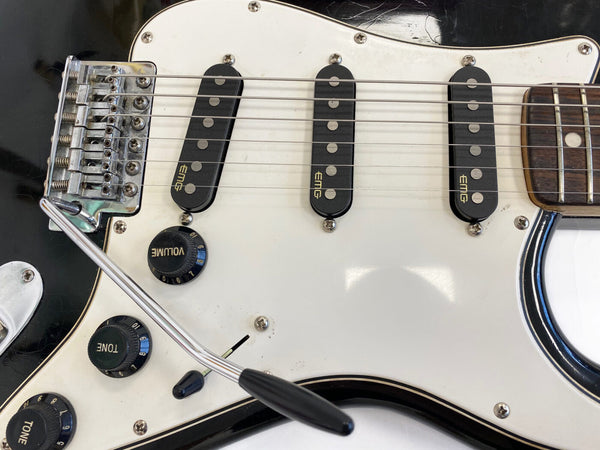 Close-up of black electric guitar with white pickguard, three EMG single-coil pickups, volume and tone control knobs, and chrome tremolo arm.