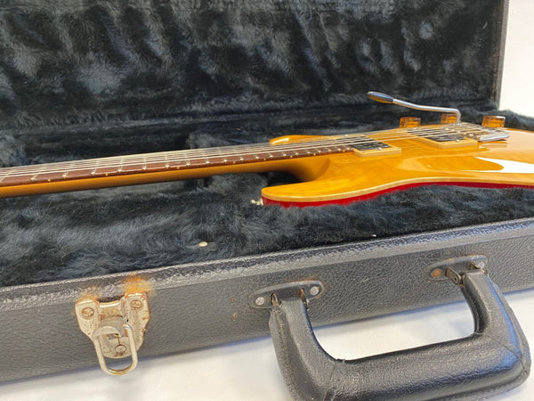 Electric guitar with a honey-colored finish and vibrato arm, placed inside a black hard case with plush interior and metal latches.