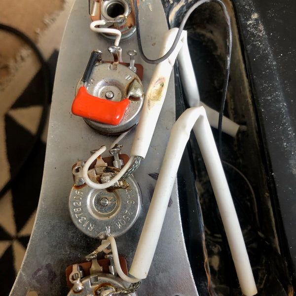 Close-up image of vintage electric guitar potentiometers with wires and capacitor on metal control plate, showing internal electronics and soldering details.