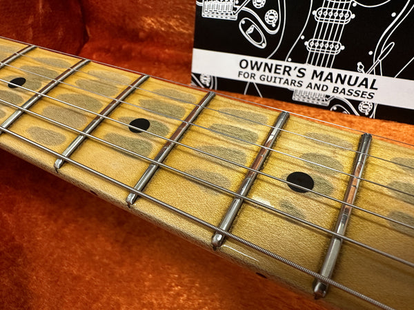 Close-up of guitar fretboard with metal frets and black dot inlays, owner’s manual for guitars and basses in the background on orange fabric surface