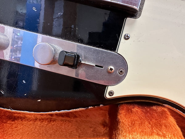 Close-up of black electric guitar control plate with chrome knob and black switch, showing wear on guitar body edge and cream pickguard, resting on orange plush guitar case lining