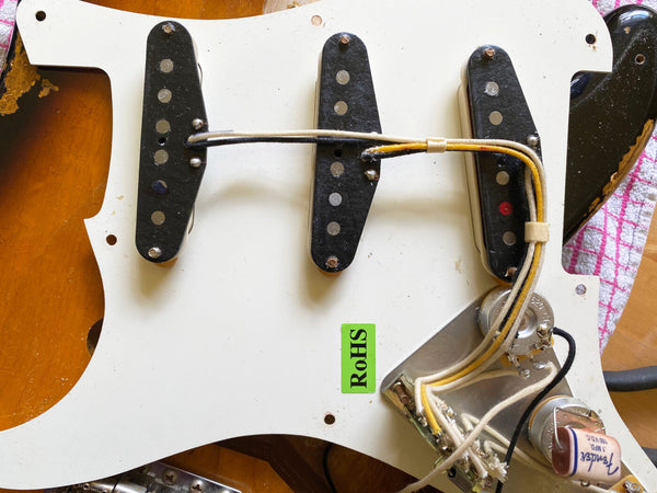 Close-up of three single coil guitar pickups mounted on a white pickguard with wiring and electronic components, labeled with a green RoHS sticker, on a sunburst electric guitar body.