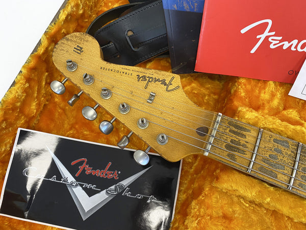 Close-up of vintage Fender Stratocaster guitar headstock with tuning pegs, original case lining, Fender Custom Shop manual, and black guitar strap on yellow plush interior.
