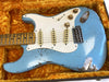 Close-up of a vintage light blue electric guitar with white pickguard and three single-coil pickups resting in an orange plush guitar case, showing worn and relic finish details on the body and fretboard.