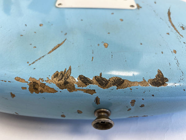 Close-up of light blue electric guitar body with heavy paint wear and chipping revealing wood beneath near strap button
